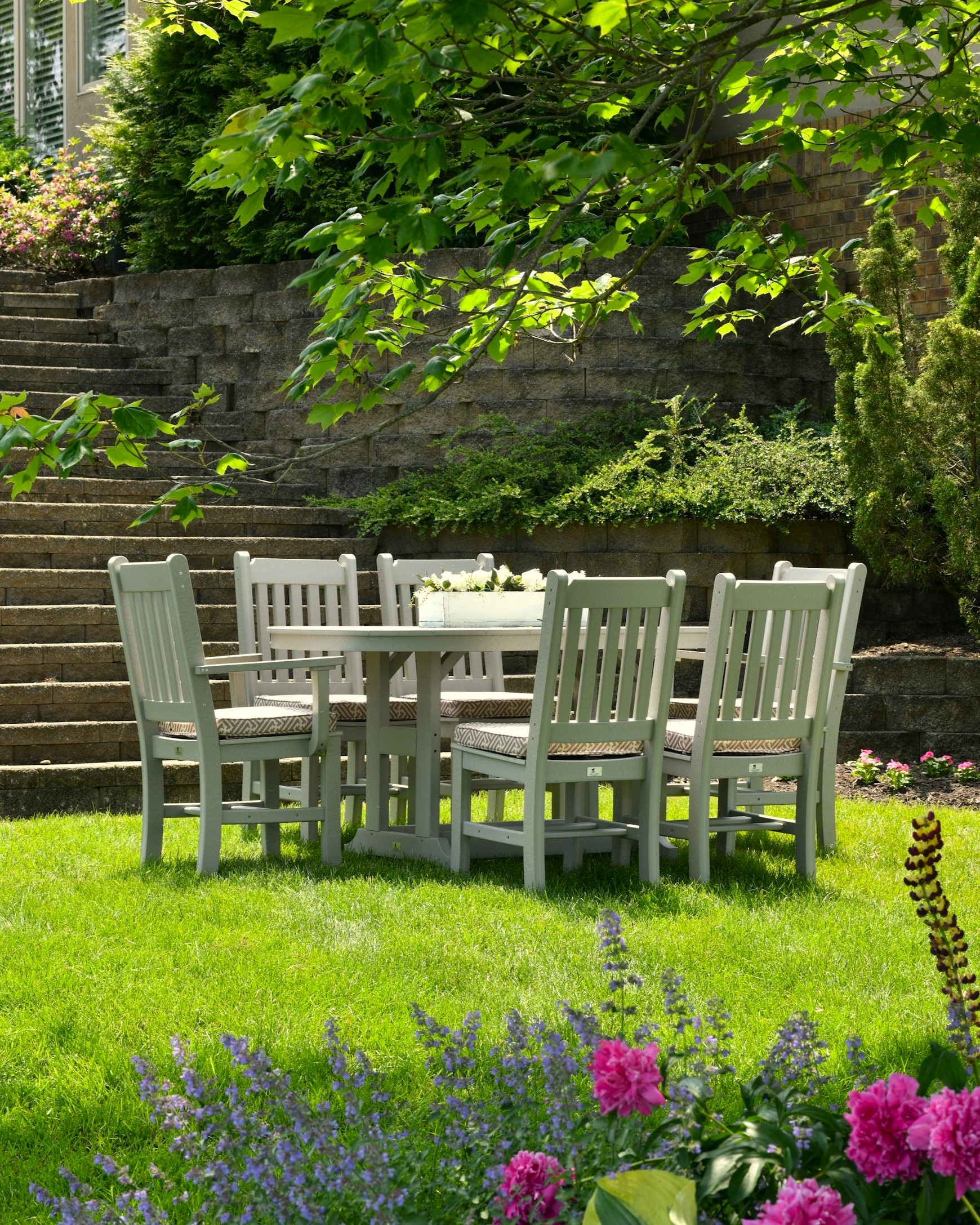Green lawn with flowers in the foreground, white square table with 6 chairs in the middle and stairs in the backround